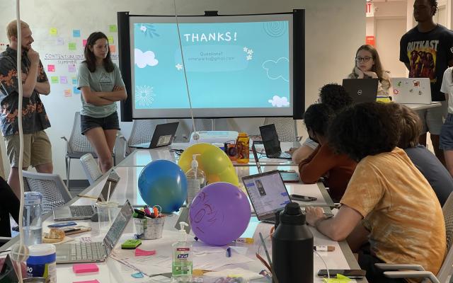 Students presenting a slide deck to a group of students at a long table filled with colorful stick notes and balloons.
