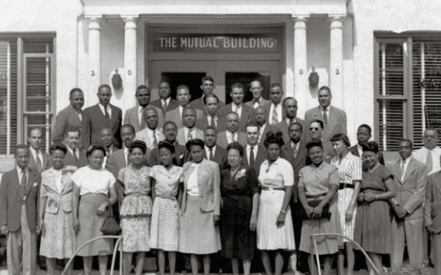 About three dozen well-dressed African American men and women posing in front of a business in the 1900s