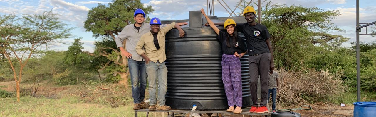 Four students posing on a platform supporting a large tank from which water is flowing into a bucket