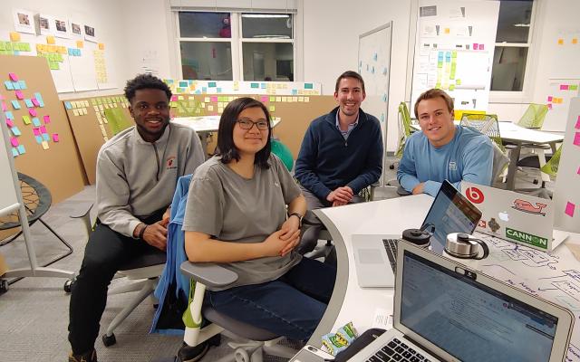 Four students smiling around white board table and walls of post-its