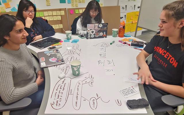 4 female students sitting around a dry-erase desk chatting about a project