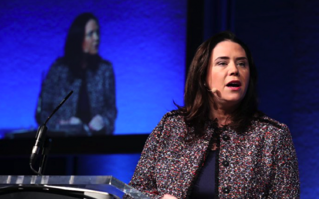 Woman with brown hair speaking at podium in front of blue background