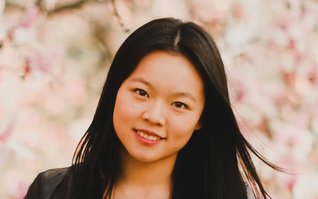 Audrey Ou, with long dark hair and wearing black is smiling with a background of pink cherry blossoms