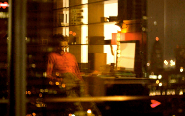 blurry shot of a man at a computer through glass with light of a city reflected.
