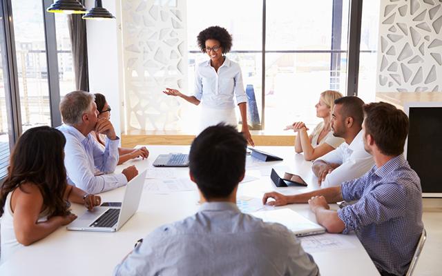 photo of people sitting around a table with a woman in front speaking